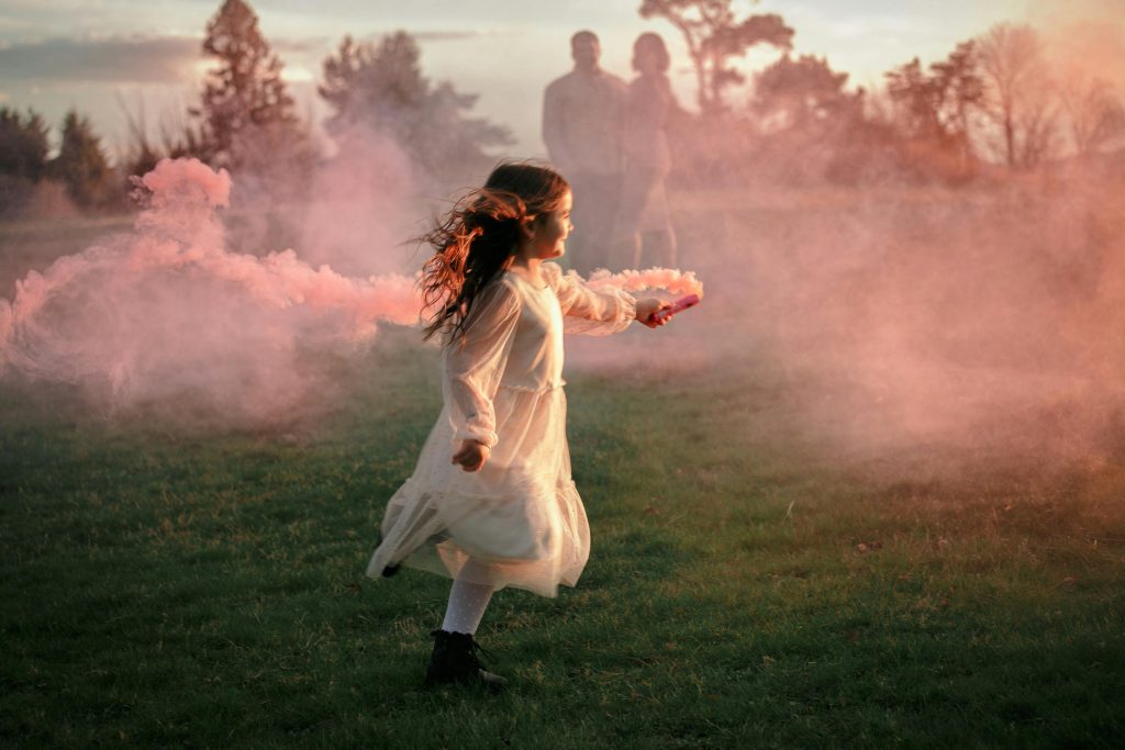 A girl in a white dress spins playfully amid pink smoke in a grassy field at sunset.